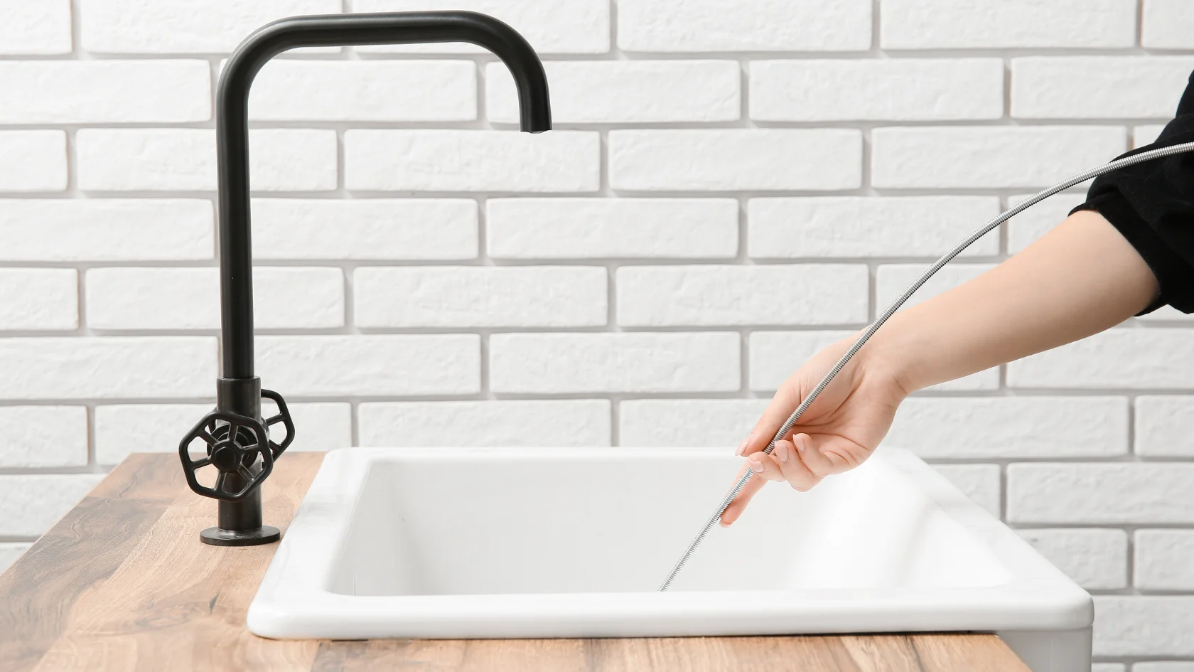 A person’s hand holds a plumbing snake, inserting it into a white sink with a black faucet, against a white tiled wall background.