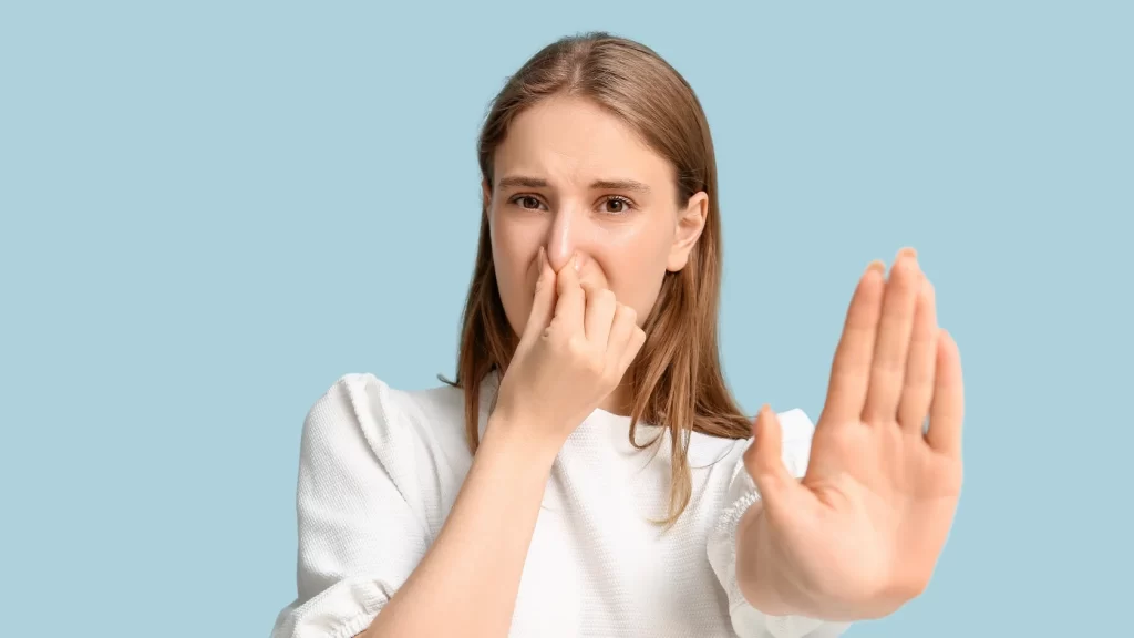 A woman holds her nose with one hand and extends her other hand forward in a stopping gesture, showing discomfort, against a light blue background.