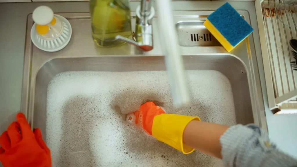 A person wearing yellow and orange rubber gloves washes dishes in a kitchen sink filled with soapy water. Dish soap, a sponge, and a drying rack are visible nearby.