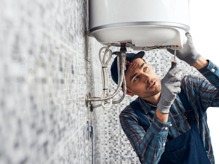 A plumber in a cap and overalls uses a screwdriver to repair a water heater mounted on a tiled wall. He is focused on his work, with wires and pipes visible—expertly handling plumbing in Toronto.