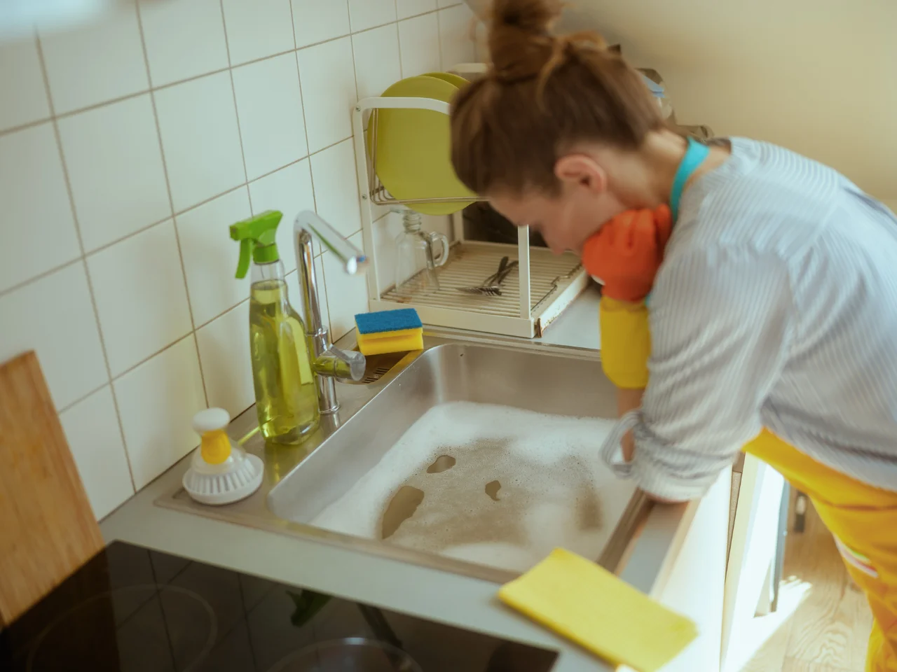 A woman wearing rubber gloves leans over a kitchen sink filled with soapy water, appearing tired or stressed, with cleaning supplies and dishes nearby.