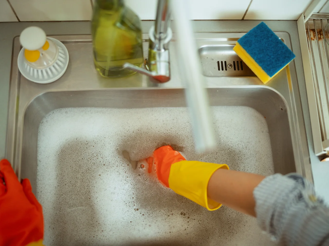 A person wearing orange and yellow rubber gloves is washing dishes in a kitchen sink filled with soapy water. A sponge and dish soap bottle are on the countertop next to the sink.