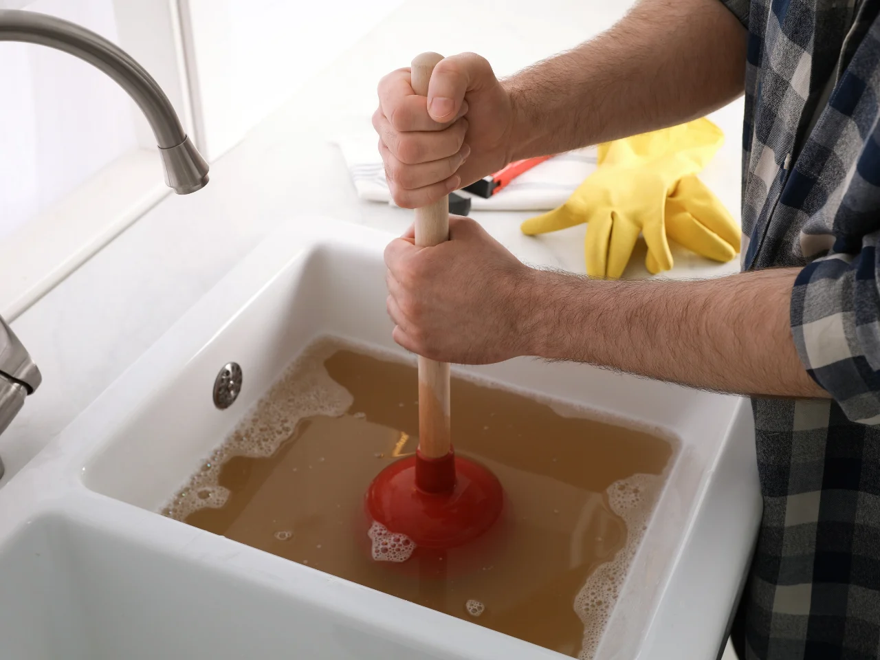 A person uses a red plunger to clear a clogged sink filled with dirty, brownish water. Yellow rubber gloves and a cleaning brush are on the counter nearby.