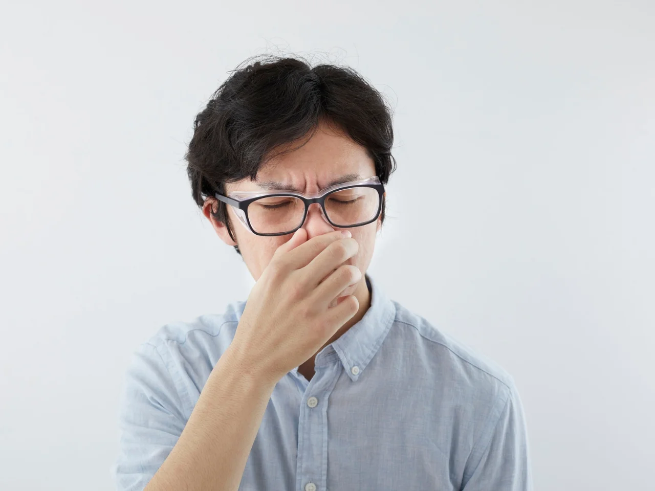 A person wearing glasses and a light blue shirt pinches their nose and closes their eyes, appearing to react to a bad smell, against a plain white background.