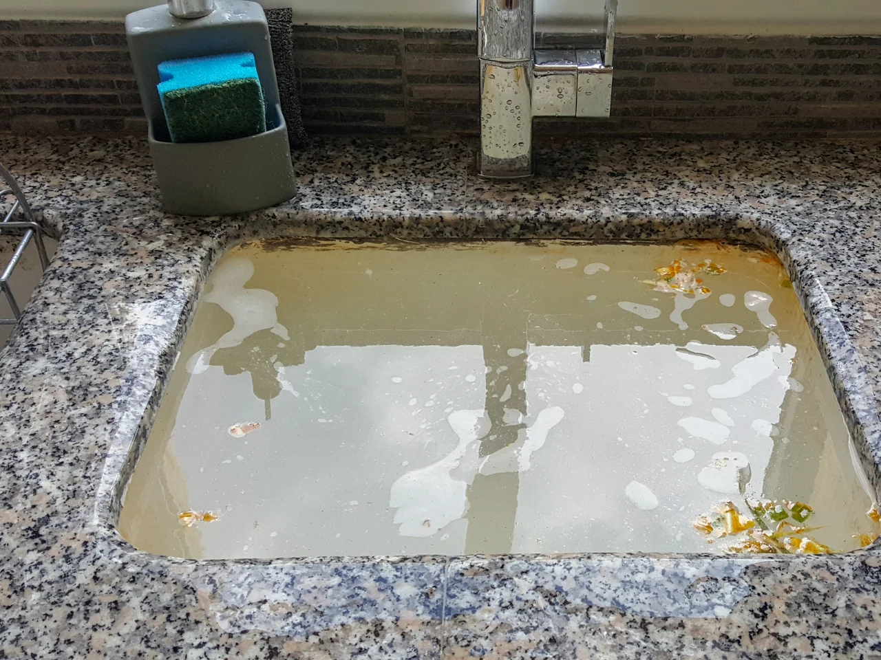 A kitchen sink filled with dirty, cloudy water and floating food debris, with a sponge and soap container on the side and water stains on the faucet.