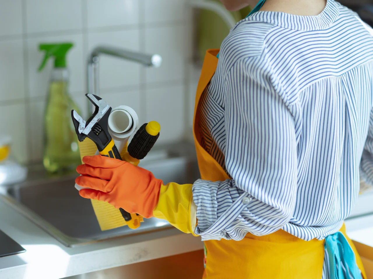 A person wearing orange gloves and an apron holds cleaning supplies and tools, including a wrench, sponge, and spray bottle, standing next to a kitchen sink.