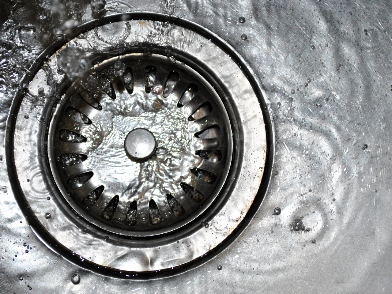 Close-up of a stainless steel kitchen sink drain with water running and visible ripples surrounding the drain.