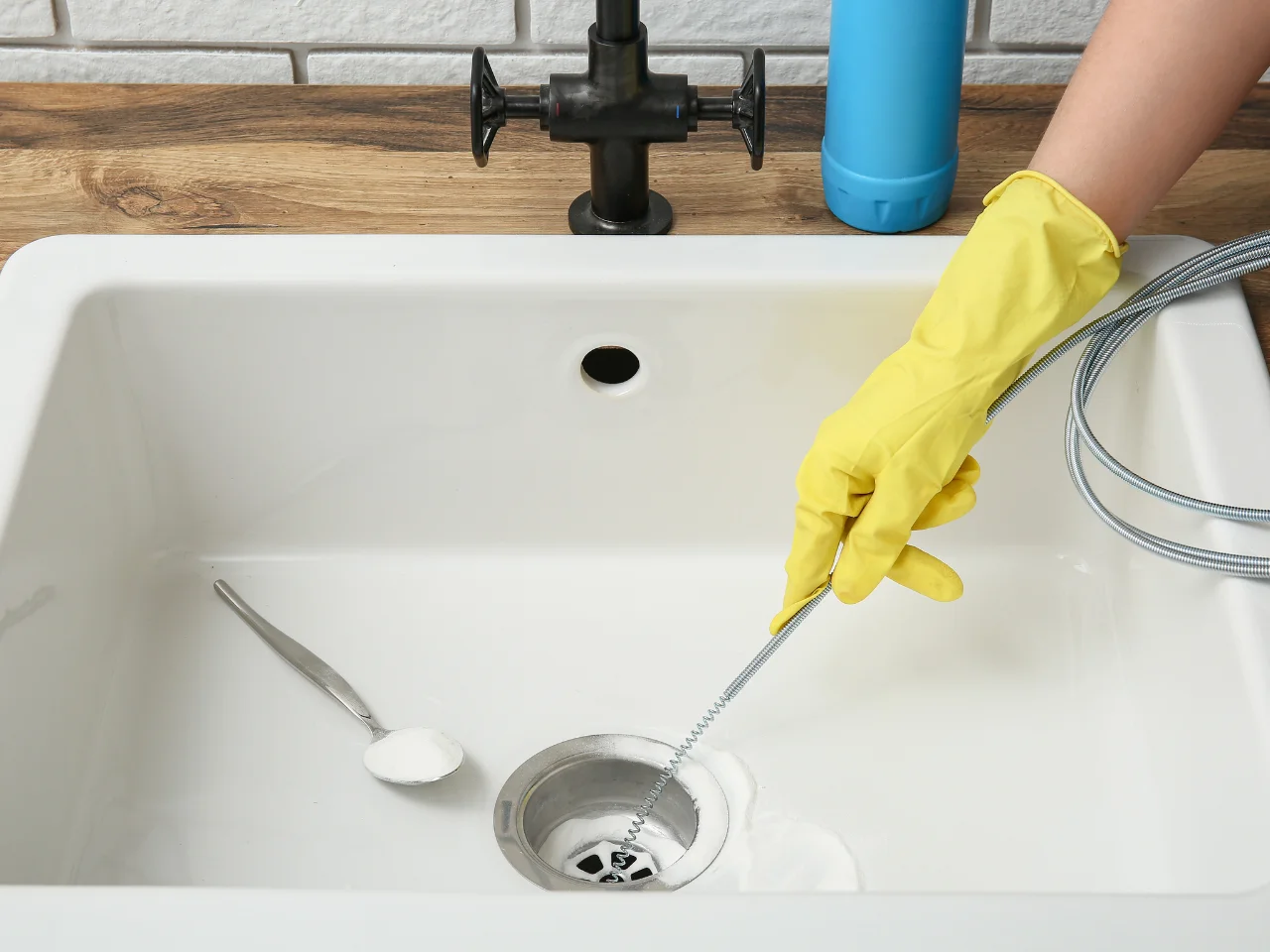 A person wearing yellow rubber gloves uses a drain snake to unclog a white sink. A spoon with baking soda and a bottle of cleaner are also visible on the sink.