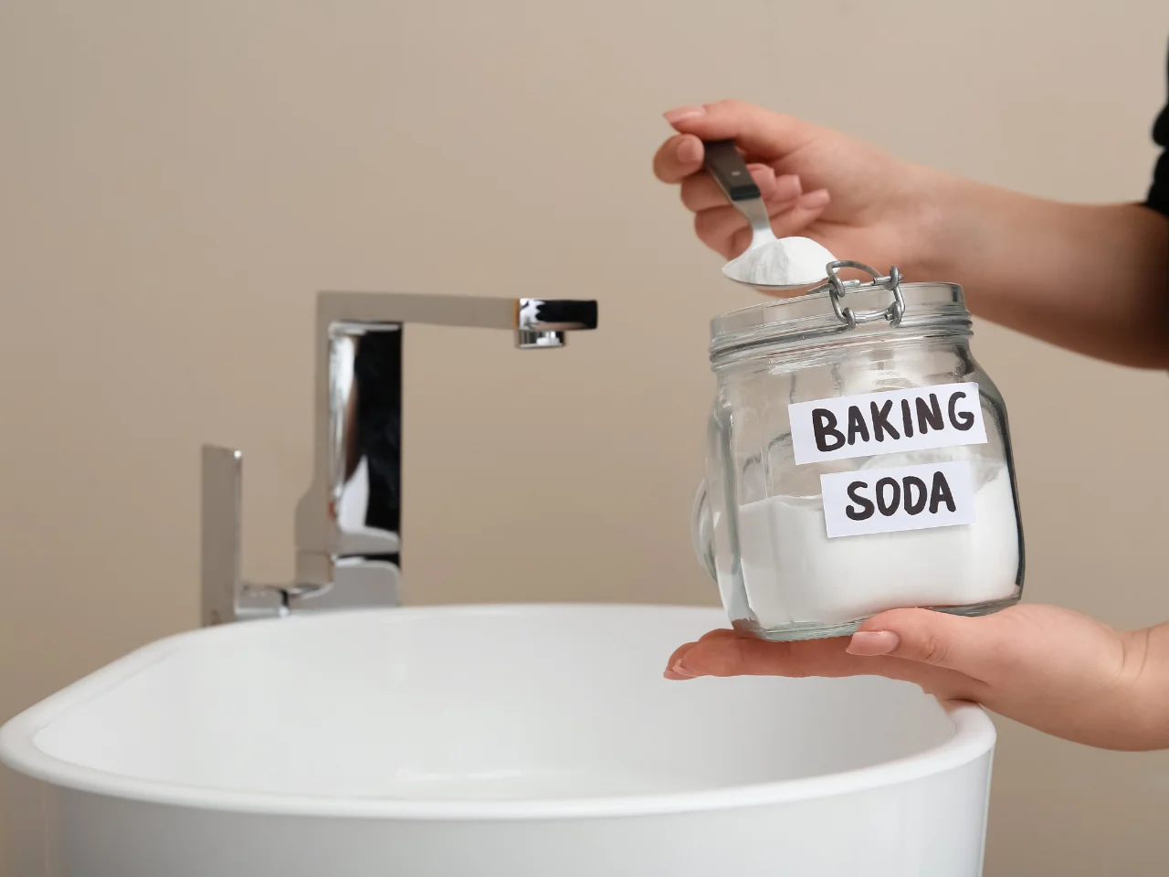 A person scoops baking soda from a labeled glass jar near a white sink and chrome faucet, with a beige wall in the background.