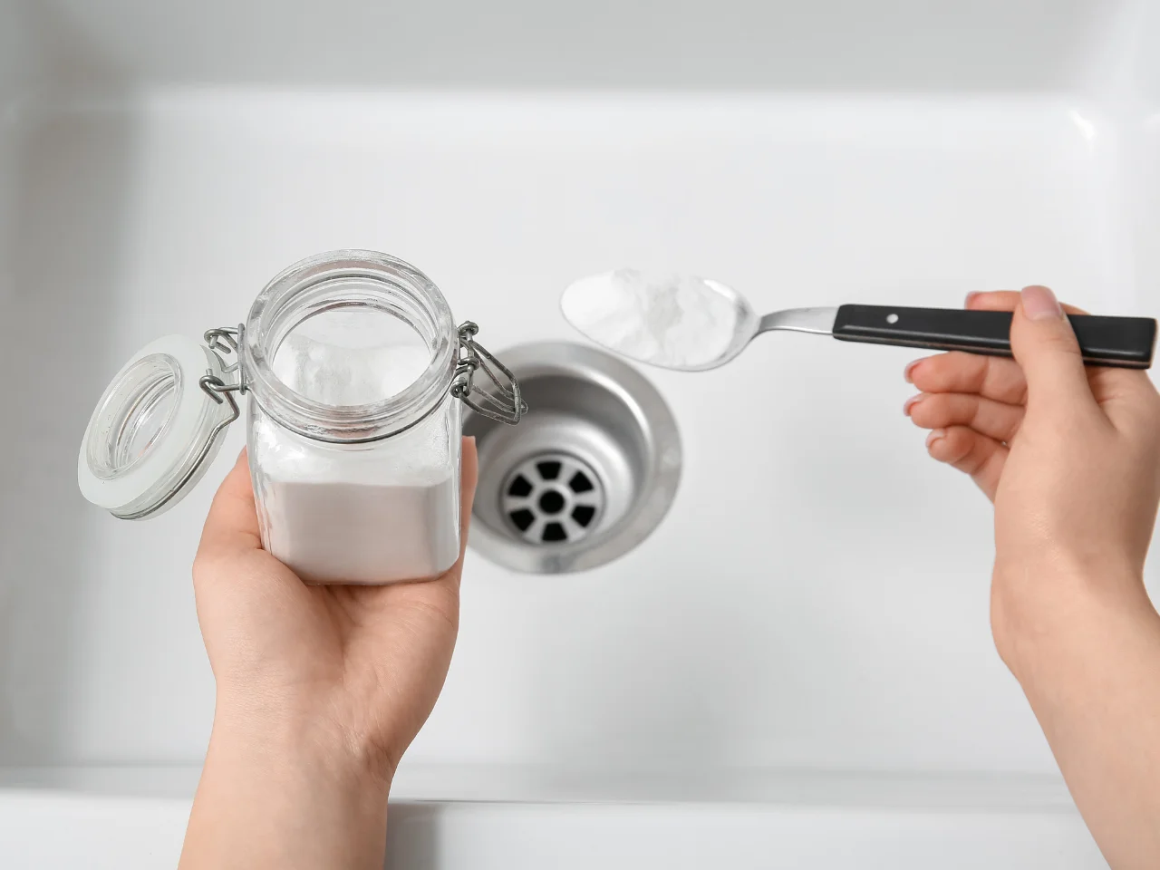 A person holds a jar of baking soda and a spoonful of it over a kitchen sink, preparing to pour the baking soda down the drain.