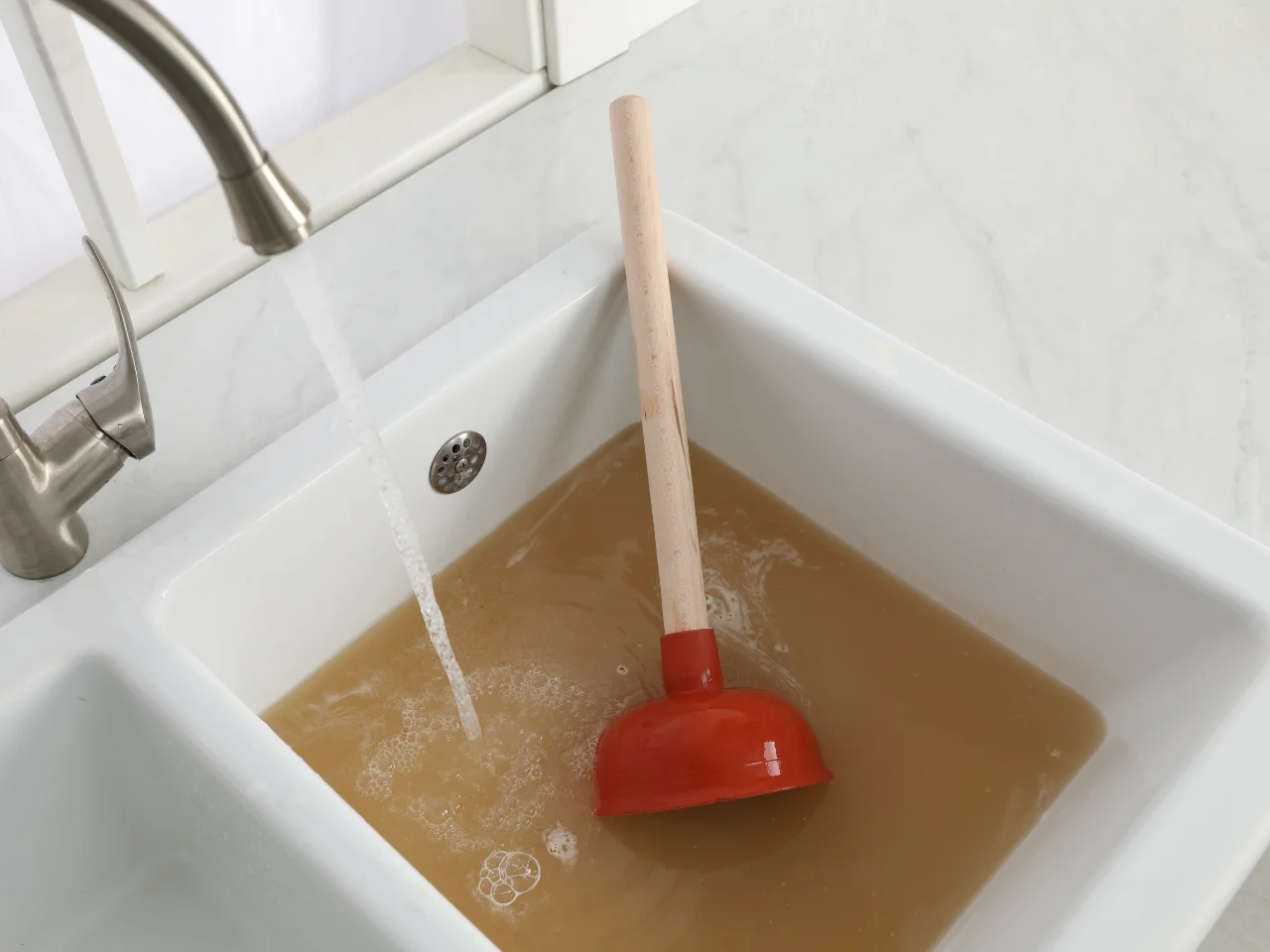 A kitchen sink filled with murky brown water, a red-handled plunger resting inside, and water running from the faucet, indicating a clogged drain.