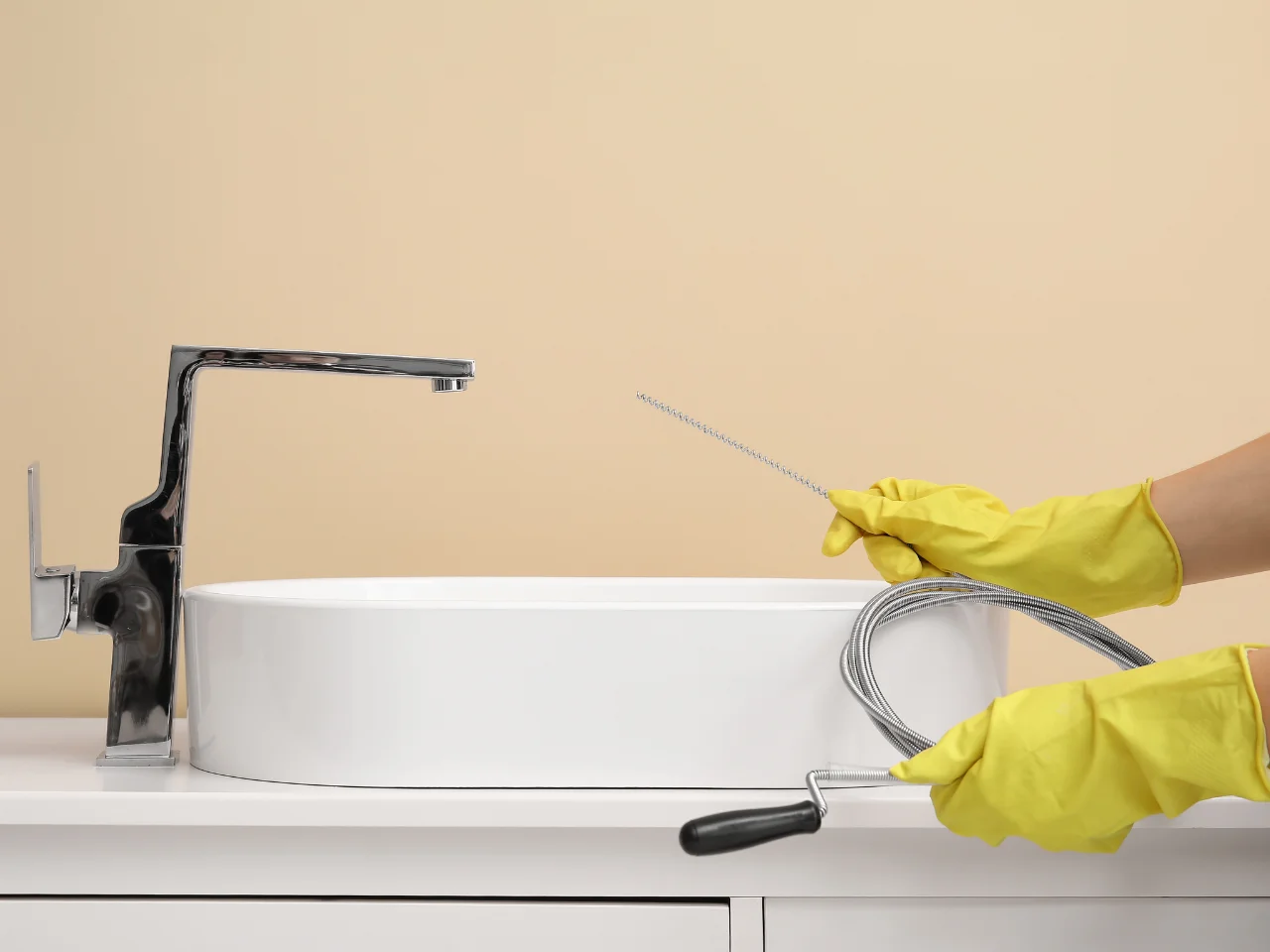 A person wearing yellow rubber gloves holds a plumbing snake tool near a modern white sink with a chrome faucet, preparing to unclog the drain. The background wall is beige.