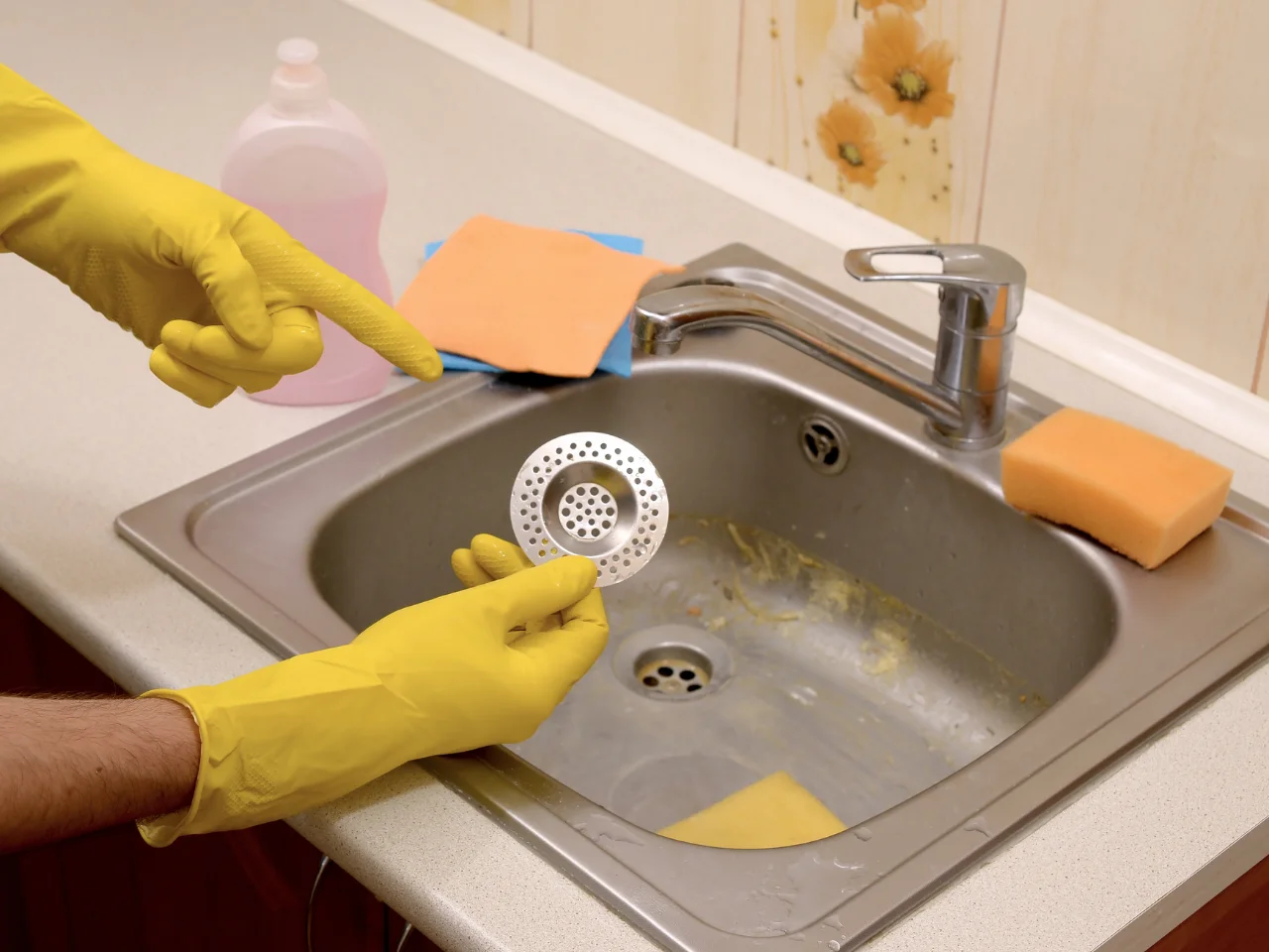 A person wearing yellow rubber gloves holds a metal sink strainer near a dirty kitchen sink with a yellow sponge, an orange cloth, a pink cleaning bottle, and floral wallpaper in the background.