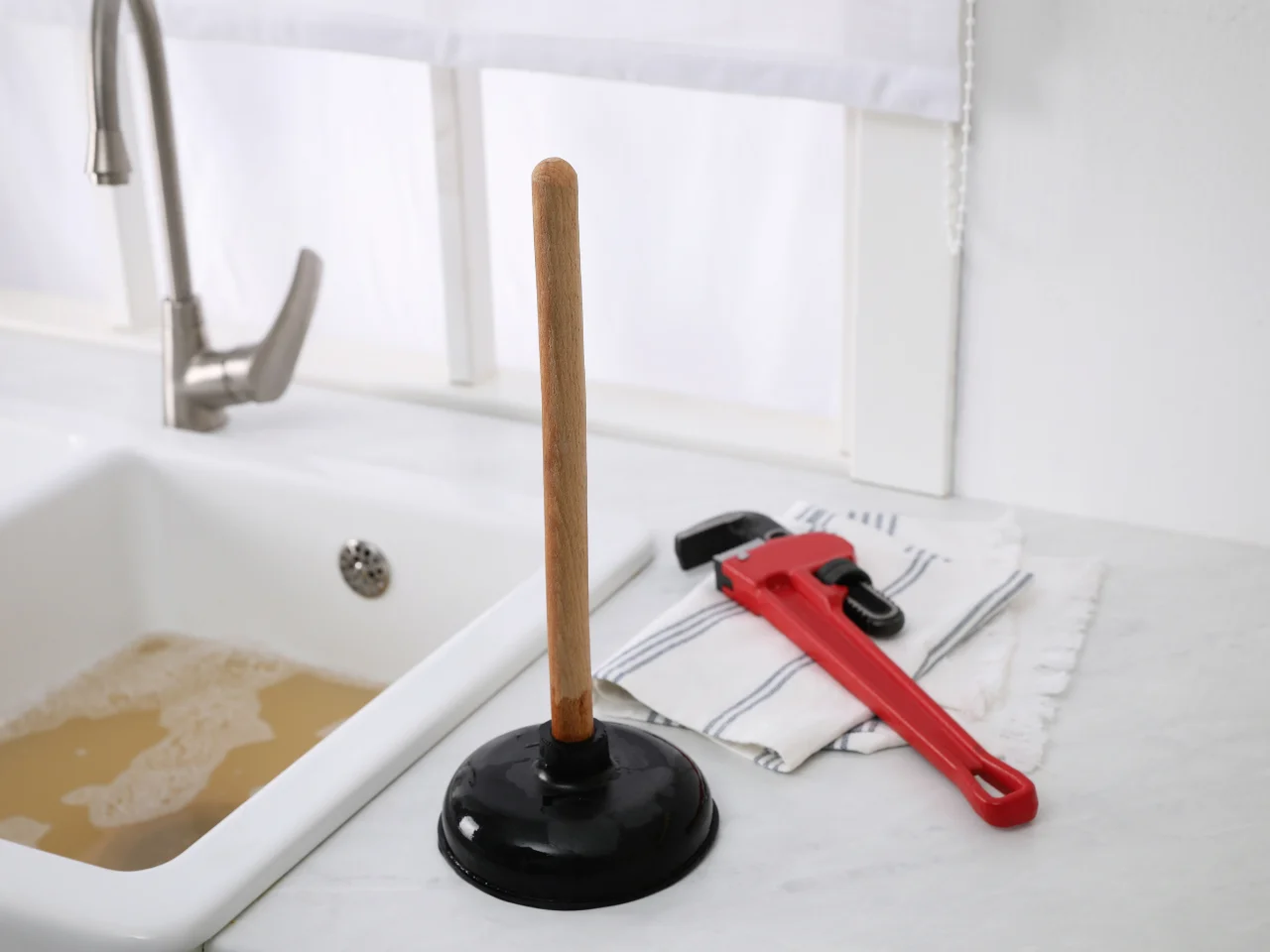 A plunger and a red pipe wrench rest on a striped cloth near a white kitchen sink filled with murky water. The background shows a faucet and a white window.