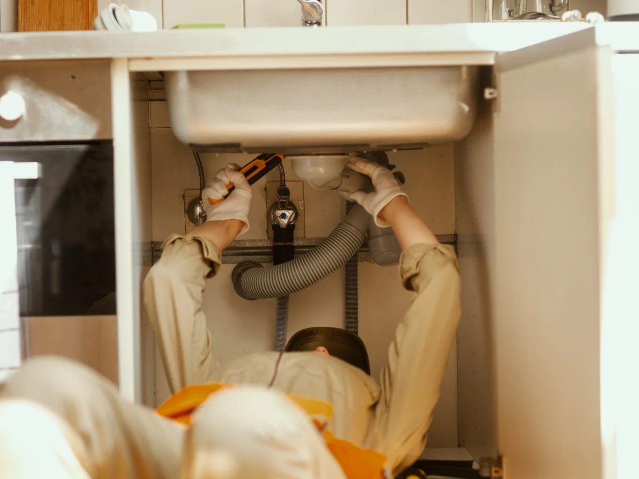 A person wearing gloves and work clothes is lying on their back under a kitchen sink, using a tool to repair plumbing inside an open cabinet.