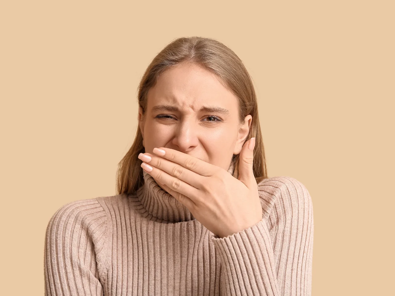 A young woman in a beige turtleneck sweater covers her mouth with her hand and looks uncomfortable or disgusted, against a plain beige background.