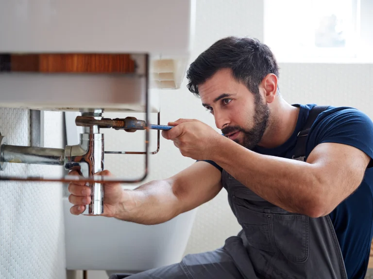 A plumber wearing overalls uses a screwdriver to fix pipes under a sink in a well-lit room, specializing in drain repair and plumbing in Toronto.