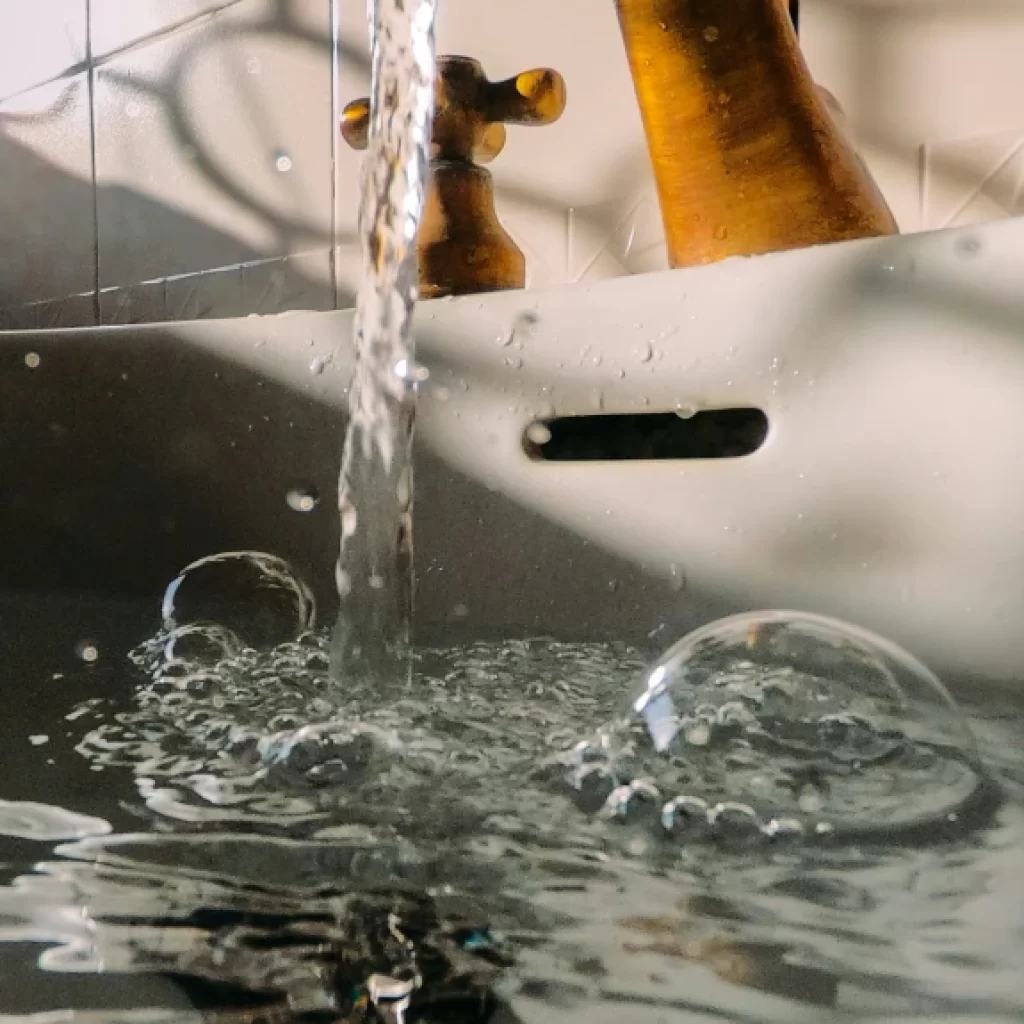 Close-up of water flowing from a tap into a white sink, creating bubbles and ripples. The brass-colored faucet and handles shine as sunlight casts shadows—perfect inspiration for drain cleaning Oakville homes.