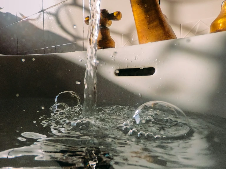 Close-up of water flowing from a tap into a white sink, creating bubbles and ripples. The brass-colored faucet and handles shine as sunlight casts shadows—perfect inspiration for drain cleaning Oakville homes.