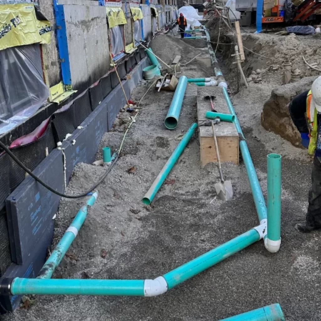 A construction worker in a helmet and safety vest levels gravel near green PVC pipes and a building foundation, highlighting drain repair Mississauga needs, with more workers and machinery in the background of the active construction site.