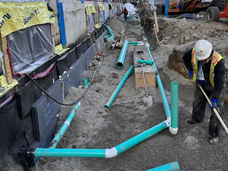 A construction worker in a helmet and safety vest levels gravel near green PVC pipes and a building foundation, highlighting drain repair Mississauga needs, with more workers and machinery in the background of the active construction site.