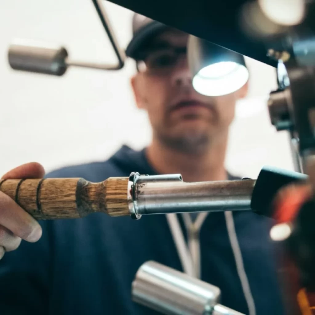 A person wearing glasses and a cap uses a wooden-handled tool to work on a metal machine, focusing intently under a bright light—a close-up capturing the precision and expertise of drain repair in Oakville.