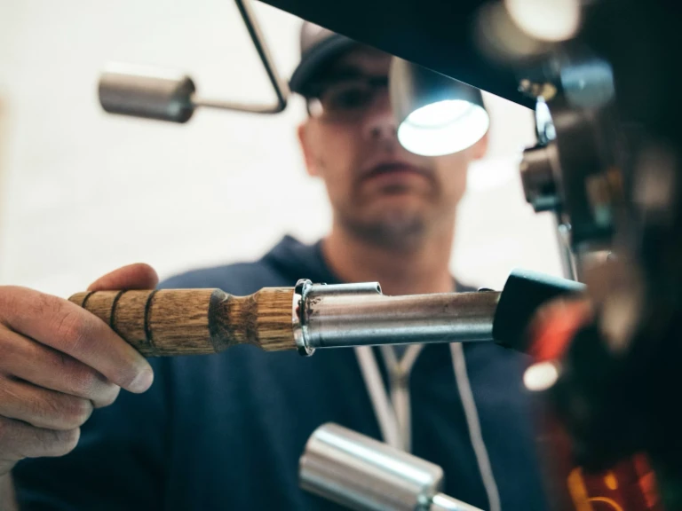 A person wearing glasses and a cap uses a wooden-handled tool to work on a metal machine, focusing intently under a bright light—a close-up capturing the precision and expertise of drain repair in Oakville.