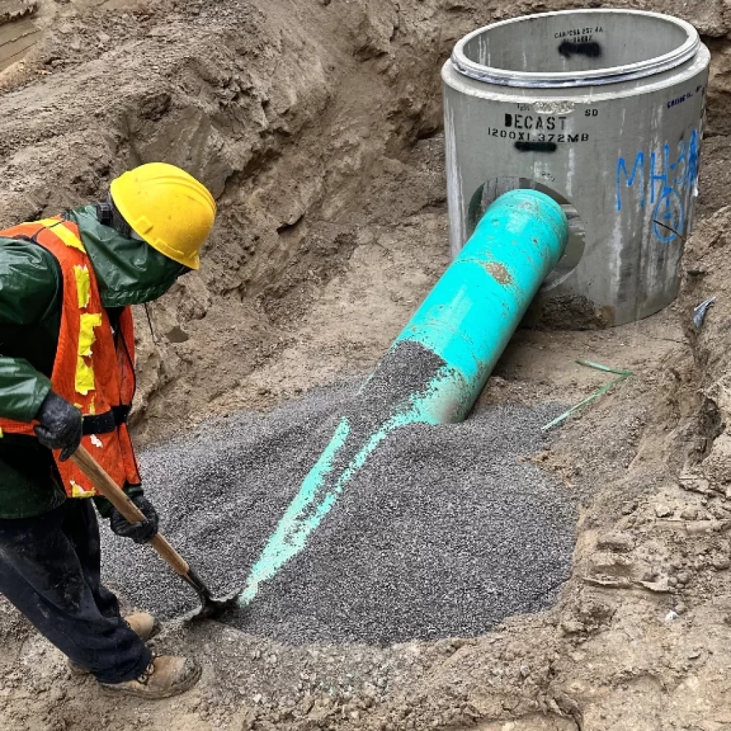 A construction worker in safety gear uses a shovel to spread gravel around a large green pipe connected to a concrete manhole during an underground drain repair Toronto installation.