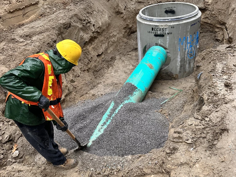 A construction worker in safety gear uses a shovel to spread gravel around a large green pipe connected to a concrete manhole during an underground drain repair Toronto installation.