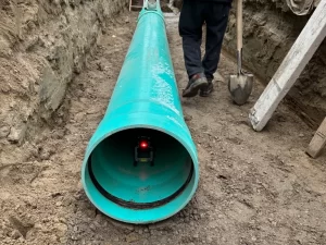 A large turquoise pipe lies in a trench, part of a drain service Mississauga project. A small device emits a red light inside the pipe, while a person with a shovel stands nearby and dirt walls line the dug-out area.