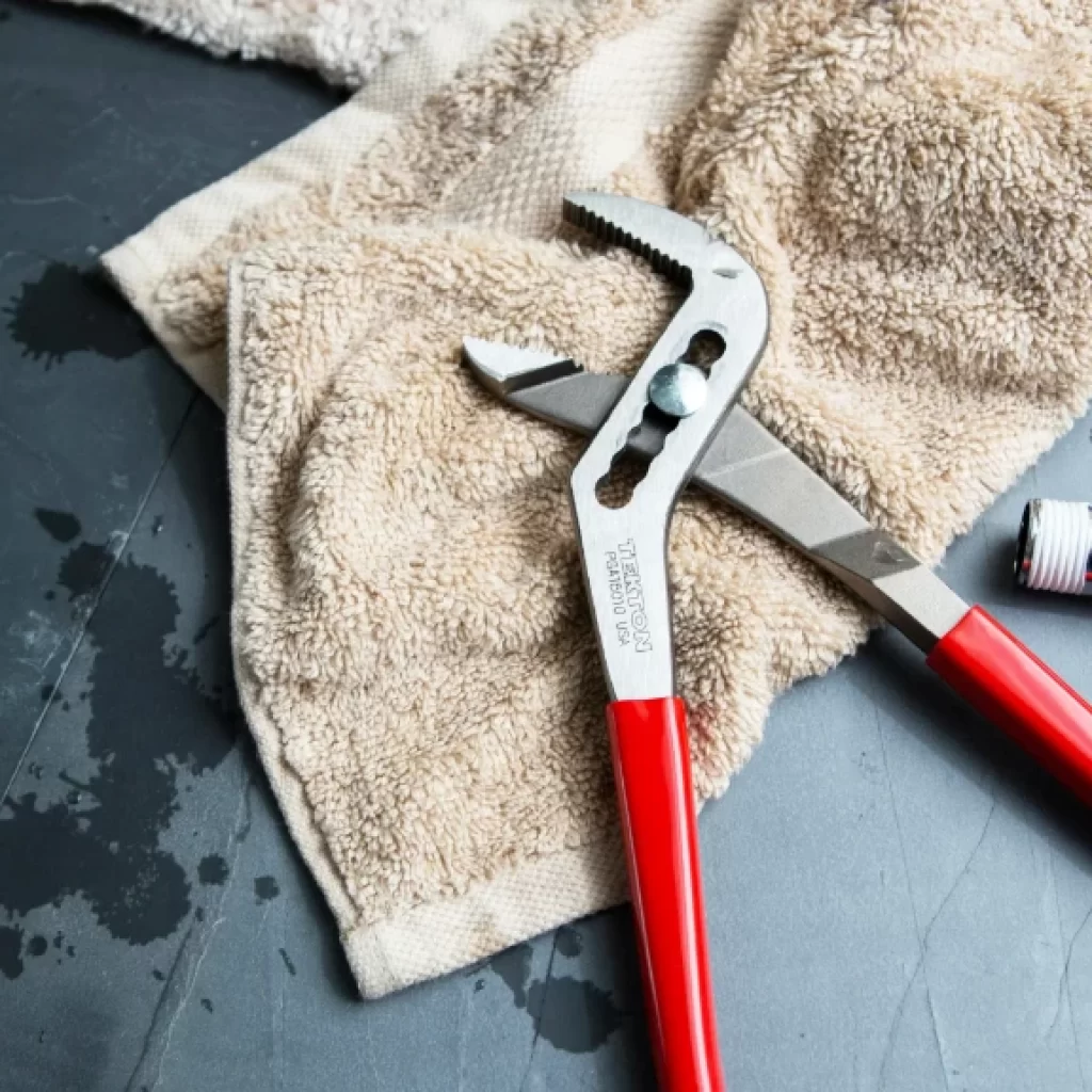 A pair of red-handled adjustable pliers and a metal pipe fitting rest on a beige towel with water drops visible on a dark surface—tools ready for an emergency plumber.