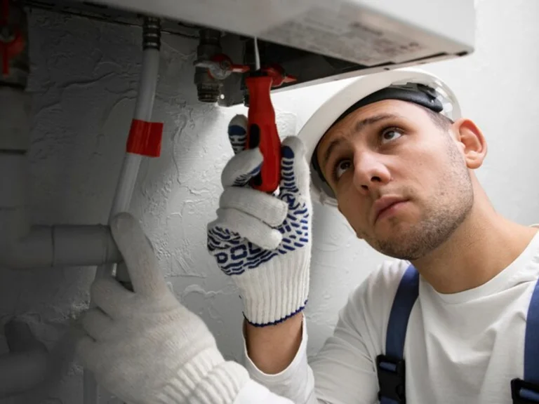A man wearing a hard hat and work gloves uses a red screwdriver to repair or inspect a pipe beneath a boiler, showcasing expert drain repair and plumbing in Toronto.