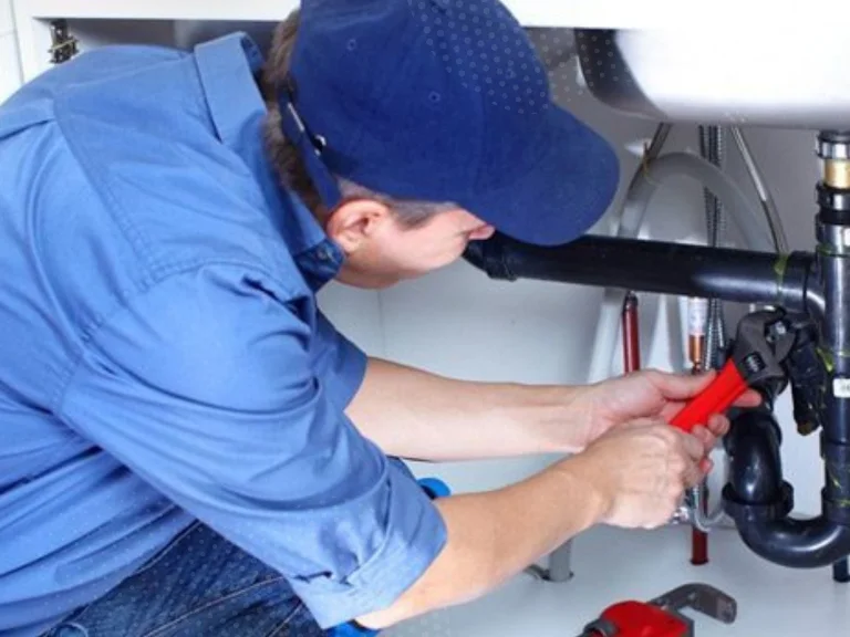 A plumber wearing a blue shirt and cap uses a red pipe wrench to fix black pipes under a sink. Various tools are visible nearby, showcasing expert drain repair and plumbing in Toronto.