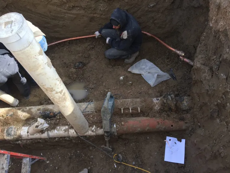 A worker in a trench tackles drain repair and plumbing in Toronto, surrounded by tools, pipes, muddy soil, and a small puddle; a sheet of paper lies near the pipes.