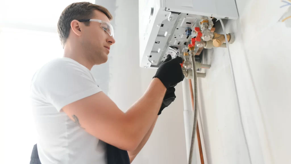 A person wearing safety glasses and gloves is servicing or installing a wall-mounted gas boiler, adjusting controls and checking pipes inside a bright room.