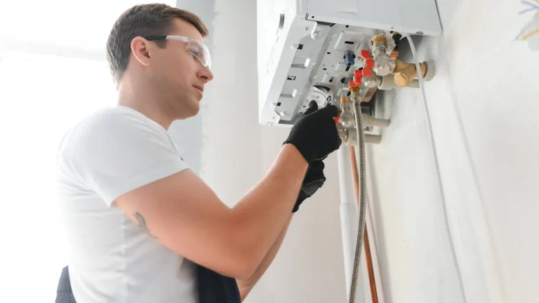 A person wearing safety glasses and gloves is servicing or installing a wall-mounted gas boiler, adjusting controls and checking pipes inside a bright room.