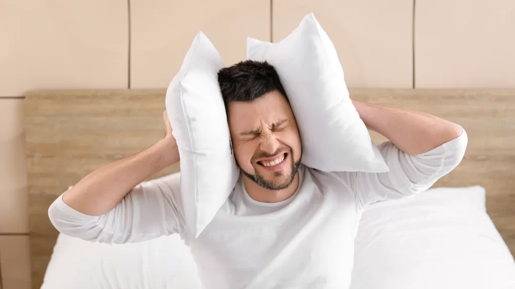A man sits on a bed pressing two pillows against his ears, eyes shut tightly and face showing frustration or discomfort, possibly disturbed by noise.
