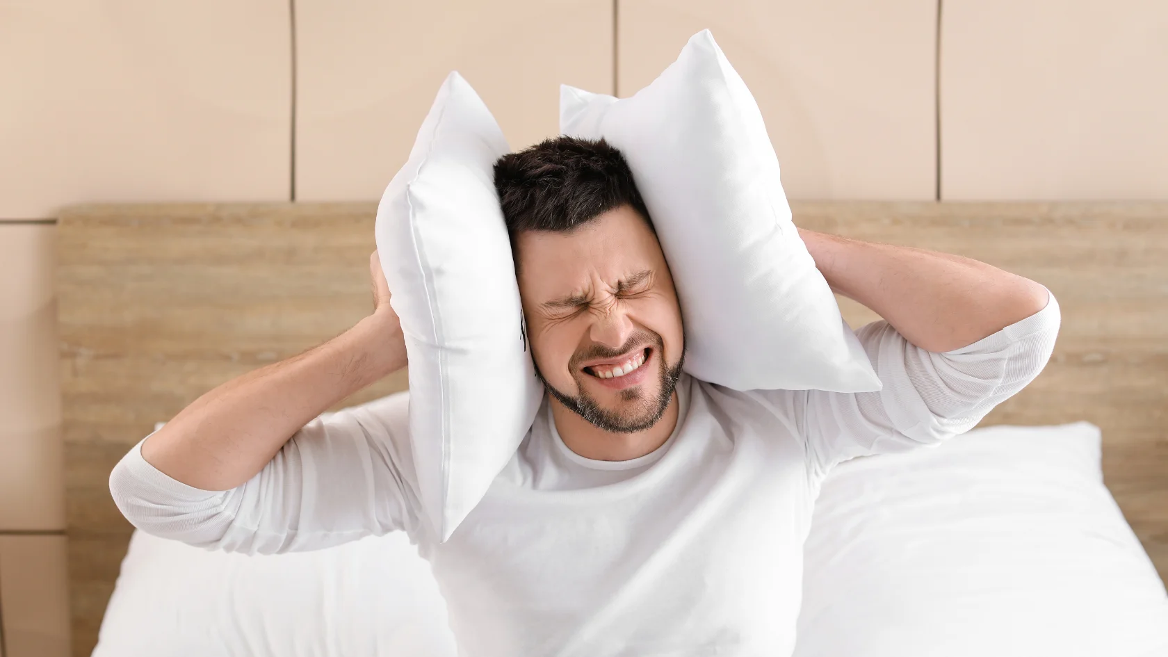 A man sits on a bed pressing two pillows against his ears, eyes shut tightly and face showing frustration or discomfort, possibly disturbed by noise.