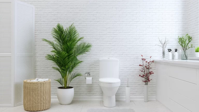 Modern bathroom with a white toilet, potted green plants, wicker laundry basket, and white brick walls. There is a white vanity with toiletries and minimalist decor on the counter. The space is bright and clean.