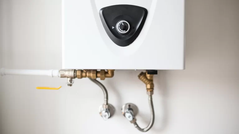 A close-up of a white tankless water heater mounted on a wall, with visible pipes and valves connected at the bottom.