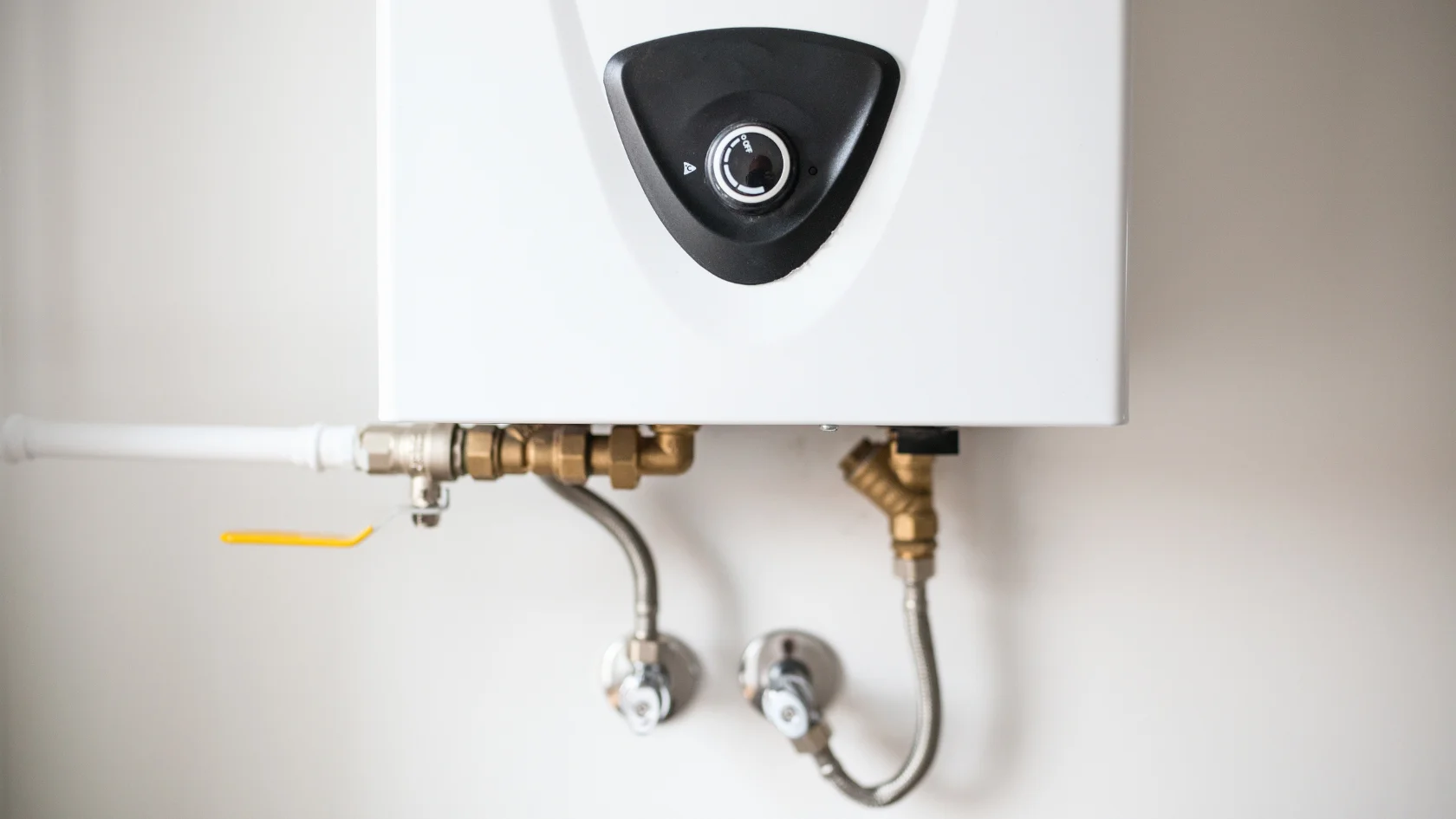 A close-up of a white tankless water heater mounted on a wall, with visible pipes and valves connected at the bottom.
