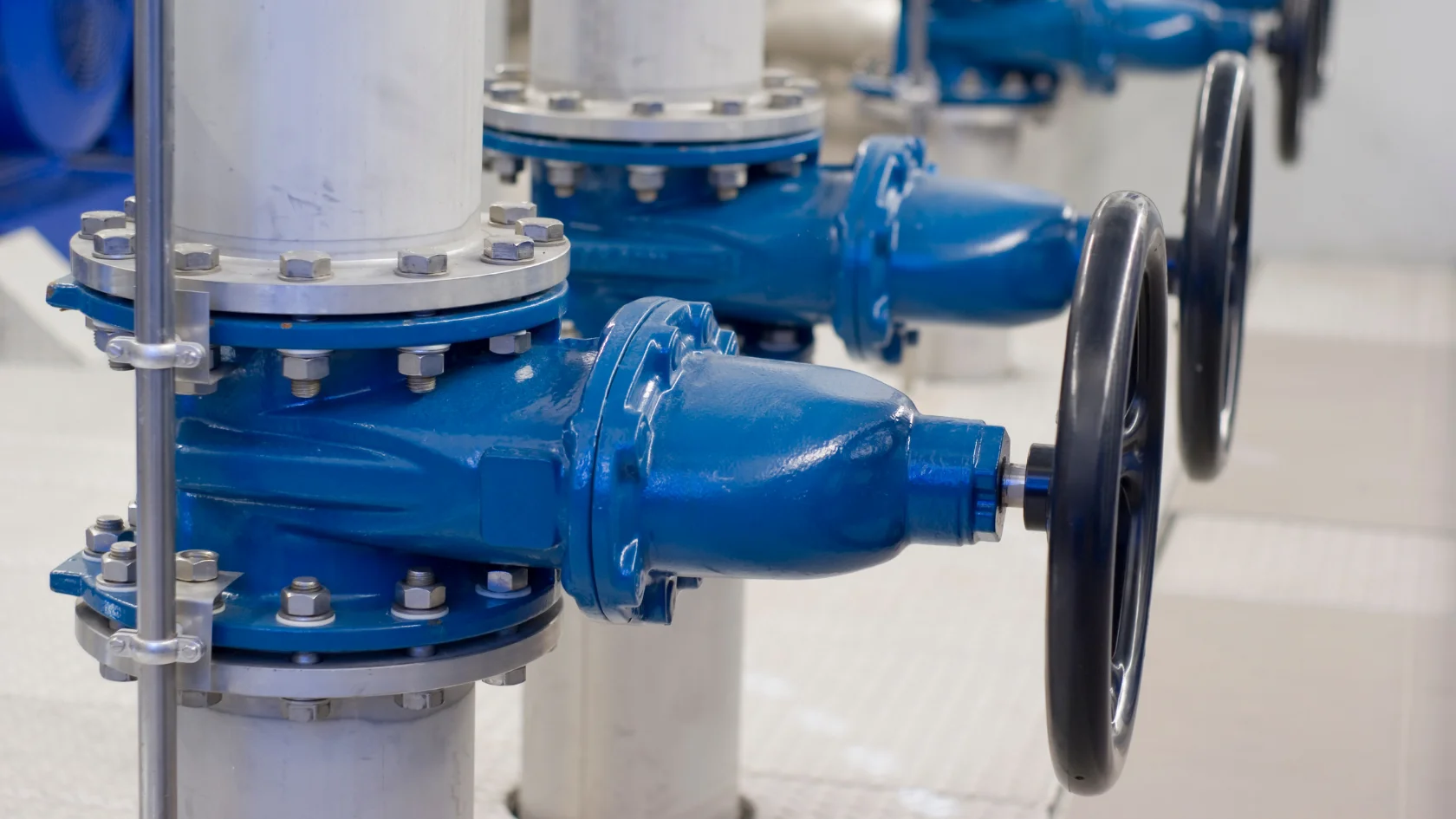 Close-up of blue industrial water valves attached to white pipes, each with a large black handwheel for manual operation, in a clean, indoor facility.