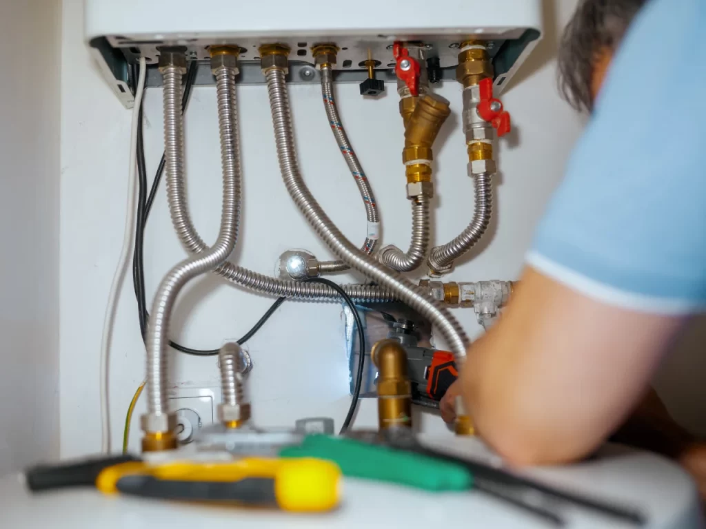 A person is repairing or installing a water heater, with several metal flexible pipes, valves, and plumbing tools visible under the unit. The persons arm is in the foreground, partially obscuring the view.