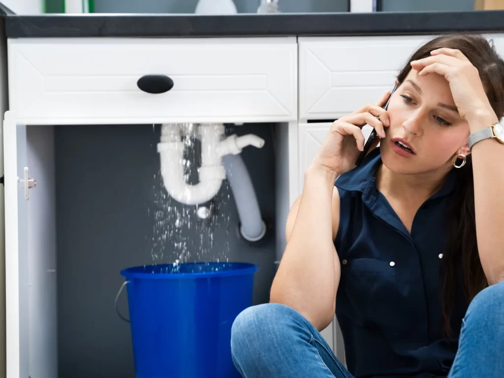 A woman sits on the floor with a worried expression, talking on the phone, while water leaks from a pipe under the sink into a blue bucket. Kitchen cabinets and plumbing are visible in the background.