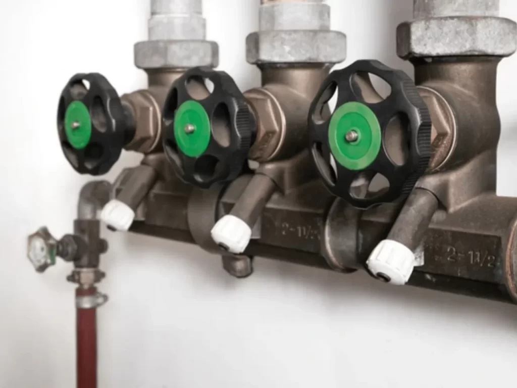 Three black and green valve handles are attached to metal pipes against a white wall, each with a white cap below the handle. The pipes appear to be part of a plumbing or heating system.