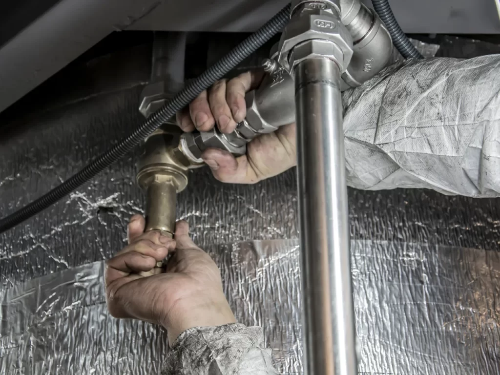 A plumber wearing a protective suit uses both hands to tighten a metal pipe joint under a sink, with silver insulation material in the background.
