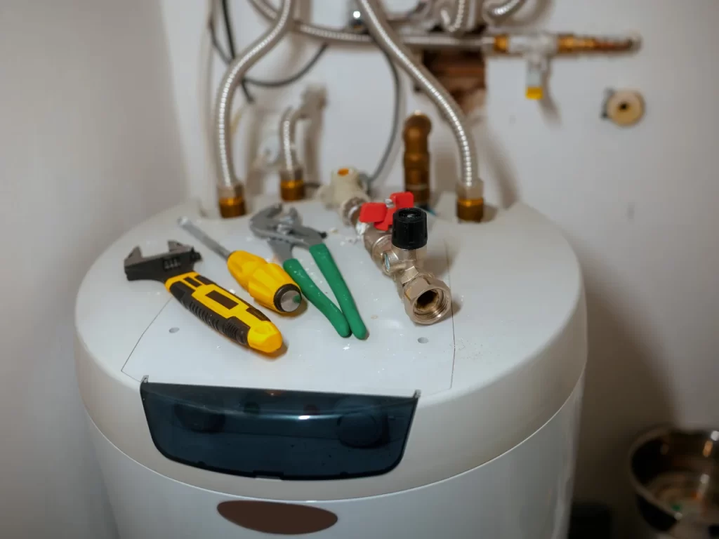 A water heater with several plumbing tools—including pliers, a wrench, a screwdriver, and a utility knife—laid out on top, with various pipes and fittings visible in the background.