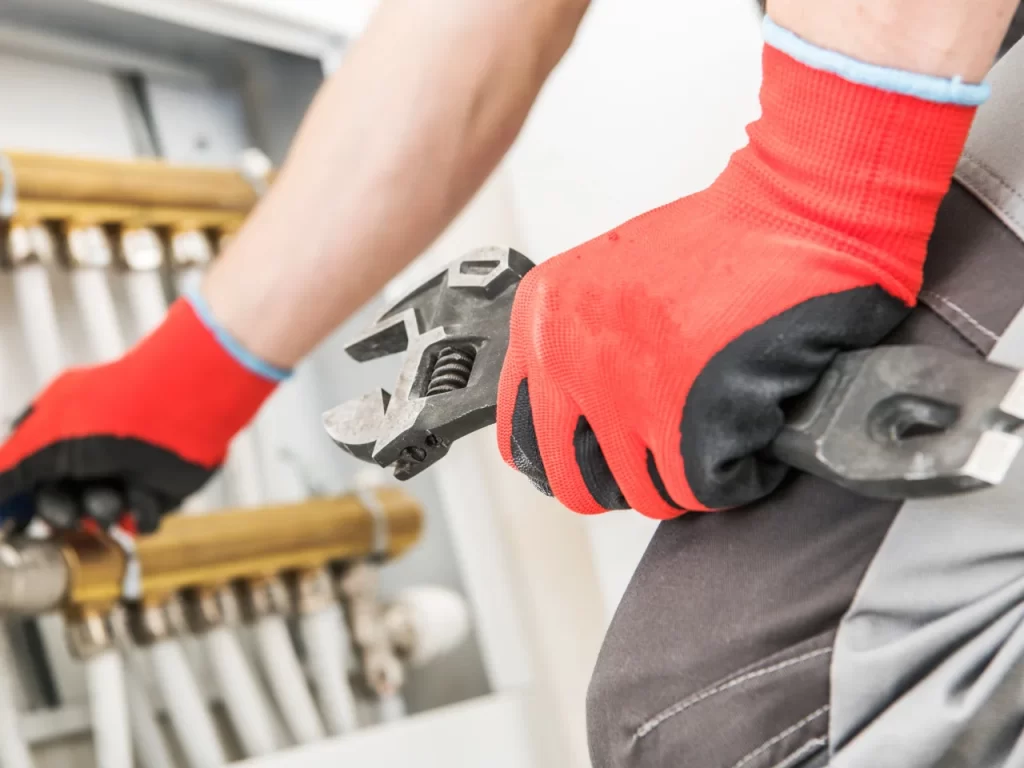 A person wearing red and black gloves uses a large adjustable wrench to work on metal pipes and valves in a utility or mechanical room.