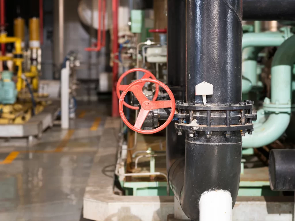 A close-up of a large black industrial pipe with a red handwheel valve in a factory or industrial facility, surrounded by machinery and equipment in the background.