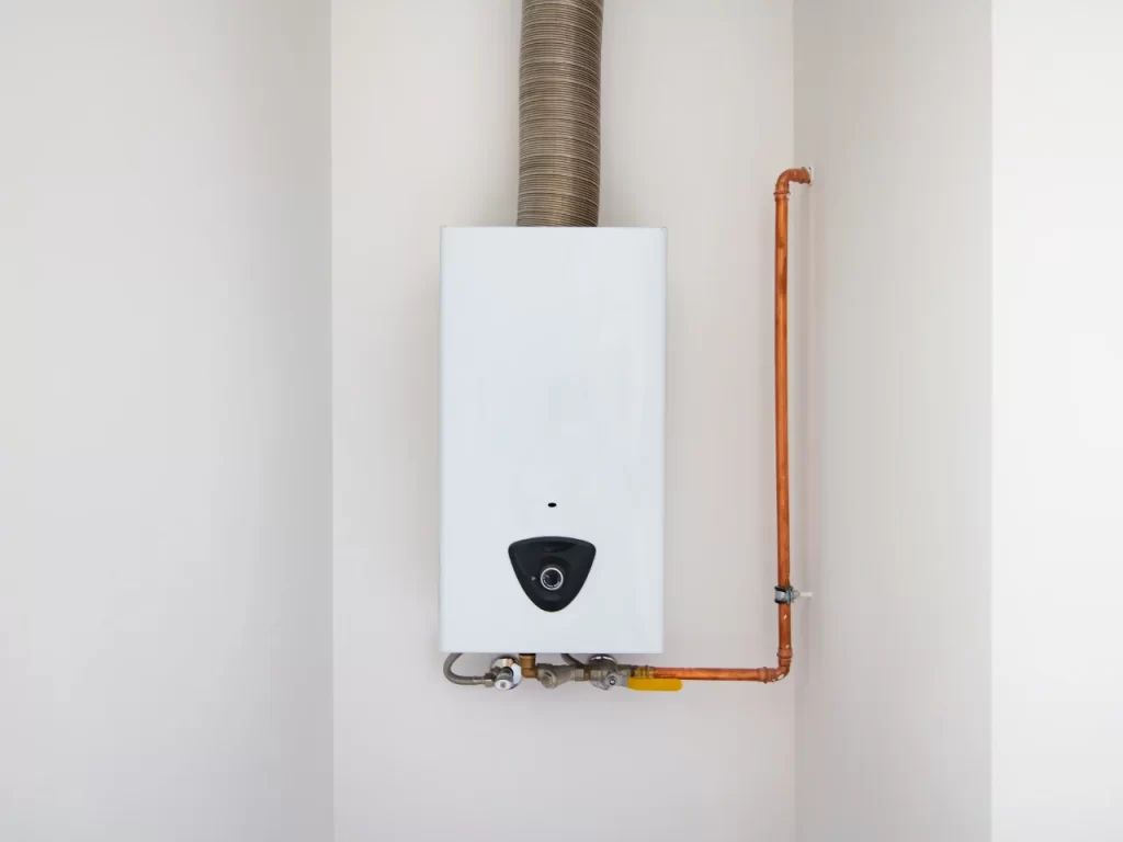 A white wall-mounted tankless water heater with a vent pipe above and copper pipes connected to the side, installed in a small, light-colored corner space.